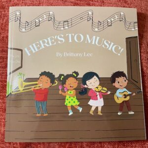 Front cover has title Here's to Music by Author Brittany Lee. There are musical notes above the title and children playing musical instruments on a stage.