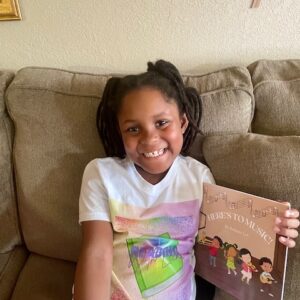 Picture includes Leena sitting on a couch with the book Here's to Music in her hand.